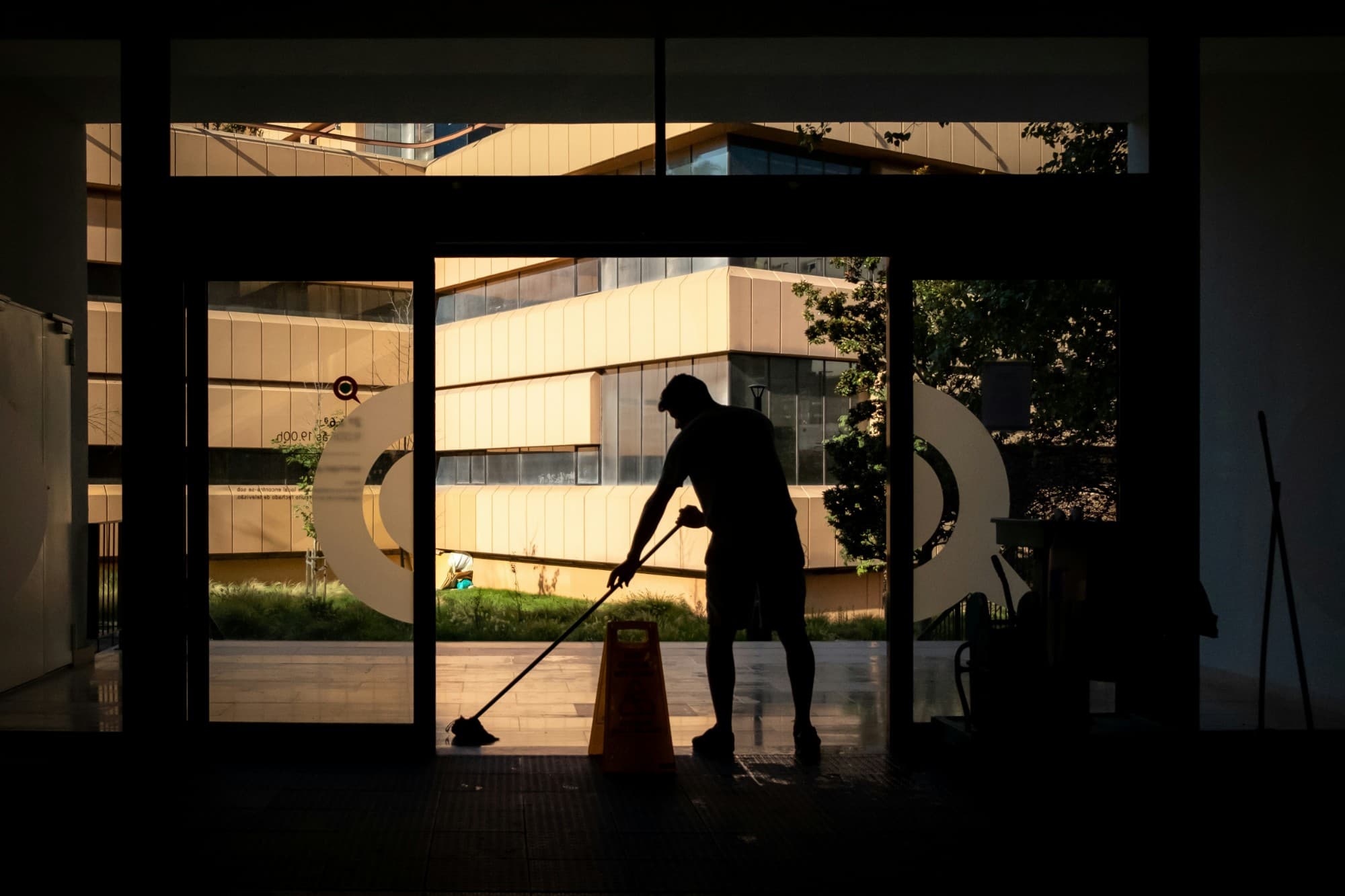 Silhouette of a cleaner mopping a building entrance beside a wet-floor sign